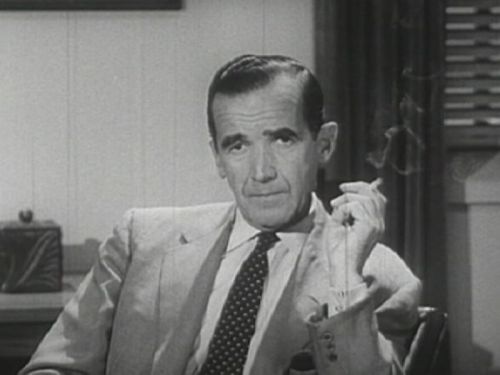 A man in a suit sits in a chair, holding a cigarette, with a serious expression in a black-and-white setting.