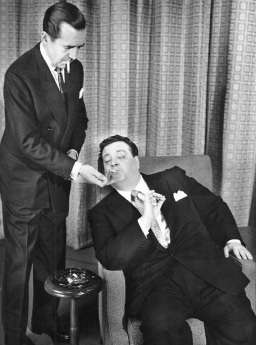 A man in a suit offers food to another man seated in a chair, both dressed formally in a vintage setting.