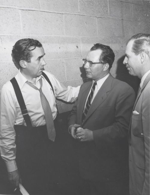 Three men in suits converse in a dimly lit hallway, one with suspenders and a tie, against a brick wall backdrop.