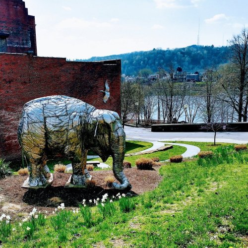 A large metallic elephant sculpture stands on a grassy area near a river, with trees and a city skyline in the background.