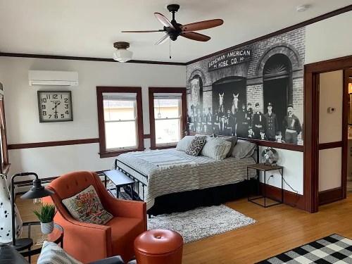 Cozy bedroom with a bed, orange armchair, and vintage wall mural featuring historical figures. Bright and inviting space.
