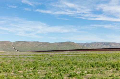 A vast landscape with green fields and a long border wall stretching across rolling hills under a blue sky.