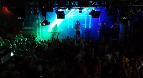 A lively concert scene with a crowd dancing, illuminated by colorful lights and a band performing on stage.