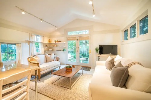 Bright, cozy living room with white sofas, a wooden coffee table, and large windows overlooking a garden.