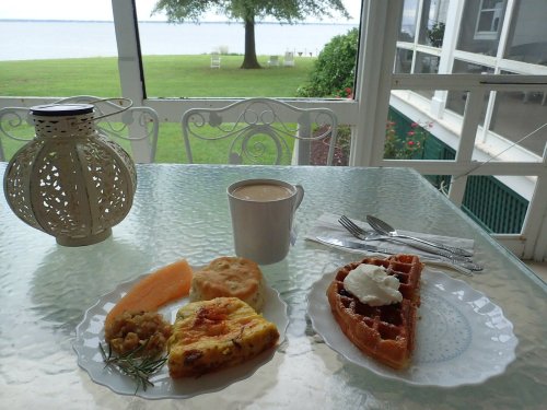 A cozy breakfast spread with a waffle, frittata, biscuit, fruit, and coffee, overlooking a serene lake view.