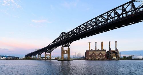 A large steel bridge spans over a river, with a power plant visible on the shore under a pastel sky.