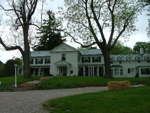 A large white house with green shutters, surrounded by trees and a grassy lawn, with a gravel path leading to the entrance.