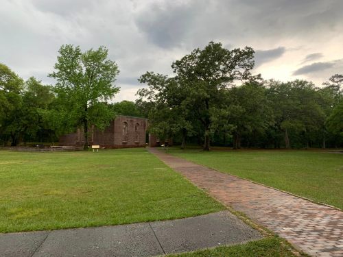 A grassy area with a pathway leading to a stone building, surrounded by trees under a cloudy sky.
