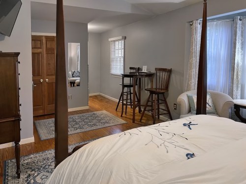 Cozy bedroom with a four-poster bed, seating area, and a small dining table, featuring warm wood tones and soft lighting.