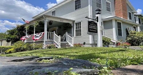 A charming guest house with a porch, American flags, and a sign, surrounded by greenery and a blue sky.