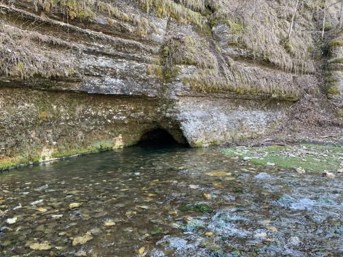 A rocky cave entrance by a clear stream, surrounded by moss-covered cliffs and scattered stones.