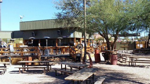 Outdoor seating area of a rustic bar with wooden tables, surrounded by trees and a sandy ground. Clear blue sky above.