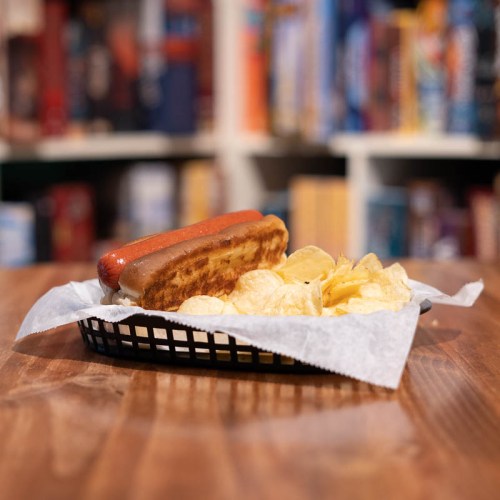 A hot dog in a basket with potato chips, set on a wooden table with blurred bookshelves in the background.