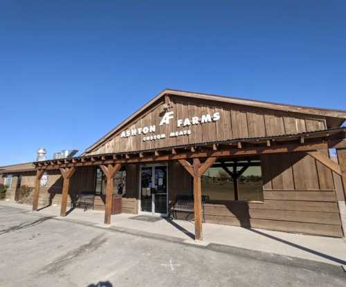 Exterior of Ashton Farms, a rustic wooden building with a sign for custom meats, under a clear blue sky.