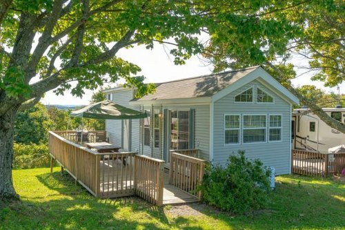 A cozy gray cottage with a wooden deck, surrounded by trees and greenery, under a clear blue sky.