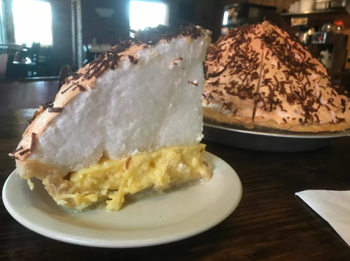 A slice of coconut cream pie on a plate, with a whole pie in the background, topped with chocolate shavings.