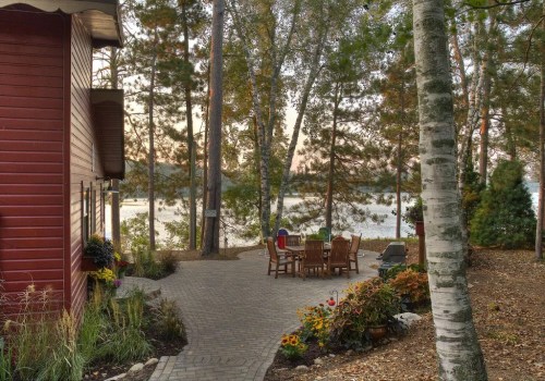 A serene lakeside patio with a stone path, surrounded by trees and flowers, featuring a table and chairs for relaxation.