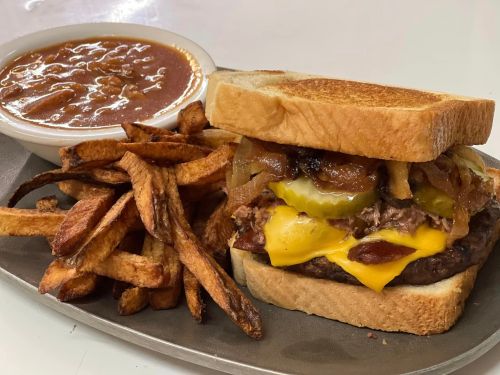 A cheeseburger with pickles and onions on toasted bread, served with fries and a bowl of chili.