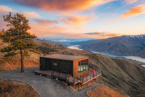 A modern cabin on a hillside with a scenic view of mountains and a river at sunset, surrounded by trees.