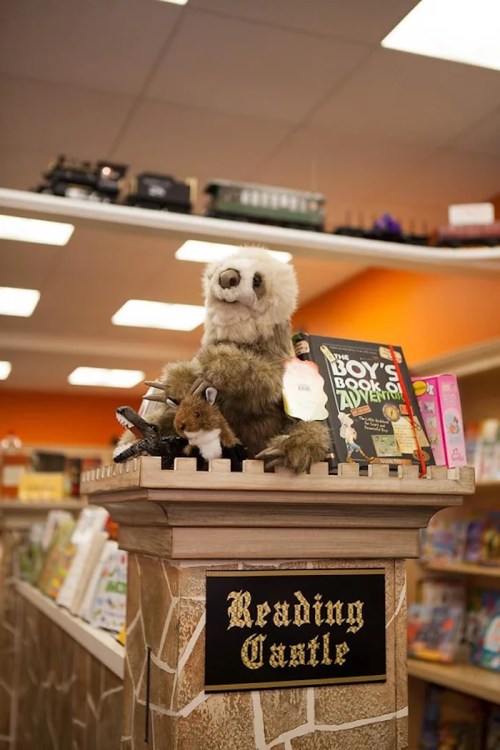 A plush animal sits atop a stone-like pedestal labeled "Reading Castle," surrounded by colorful books in a cozy bookstore.