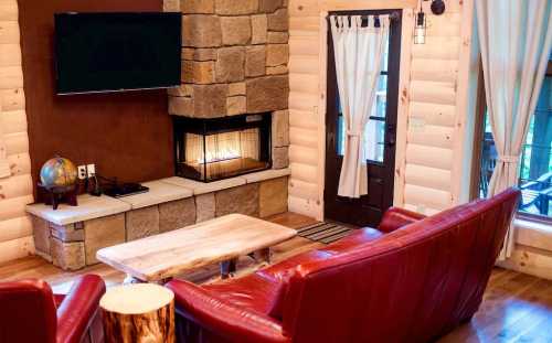 Cozy living room with a stone fireplace, red leather sofas, a wooden coffee table, and a door leading outside.