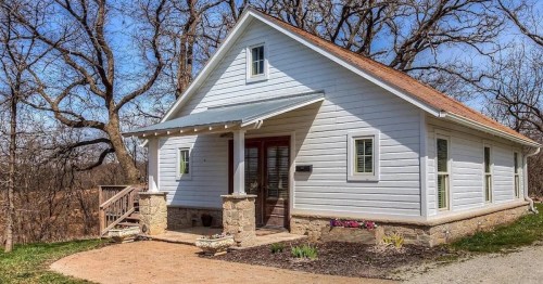 A charming white cottage with a stone porch, surrounded by trees and a clear blue sky.
