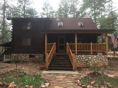 A rustic log cabin with a stone foundation, wooden porch, and an American flag, surrounded by tall pine trees.