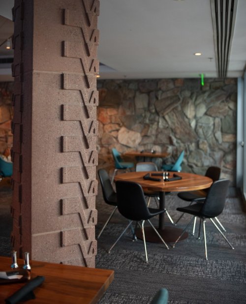 Interior of a modern restaurant featuring stone walls, wooden tables, and stylish black chairs.