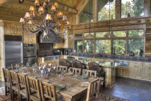 A spacious wooden kitchen with a large dining table, chandelier, and large windows overlooking a forested area.