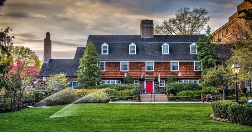 A charming two-story house with a red door, surrounded by lush greenery and a well-maintained lawn under a cloudy sky.