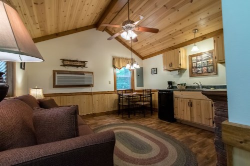 Cozy cabin interior with wooden walls, a sofa, dining table, and kitchen area, featuring warm lighting and rustic decor.