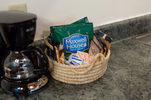 A coffee maker next to a basket filled with Maxwell House coffee packets and sugar packets on a countertop.