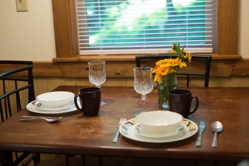 A dining table set with two plates, cups, and a vase of flowers, with sunlight streaming through a window.