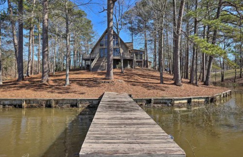 A wooden dock leads to a lakeside A-frame house surrounded by tall trees and a clear blue sky.