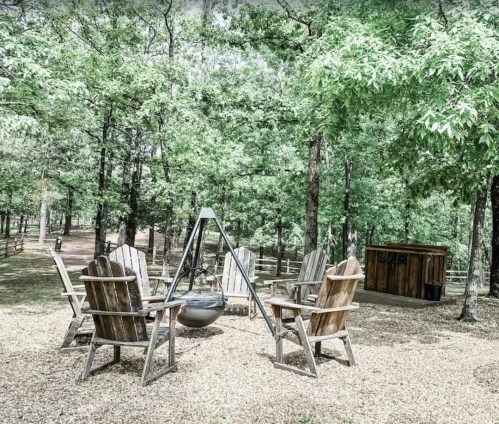 A serene outdoor space with wooden chairs arranged around a fire pit, surrounded by lush green trees.