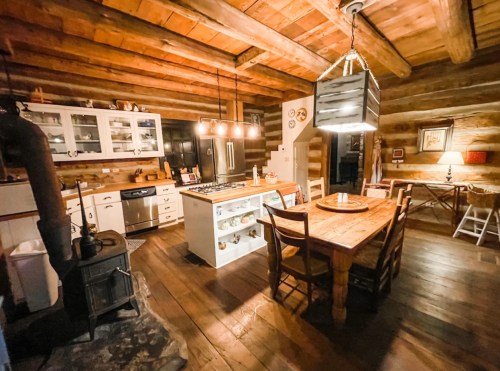 Cozy log cabin kitchen with wooden beams, a dining table, modern appliances, and warm lighting.