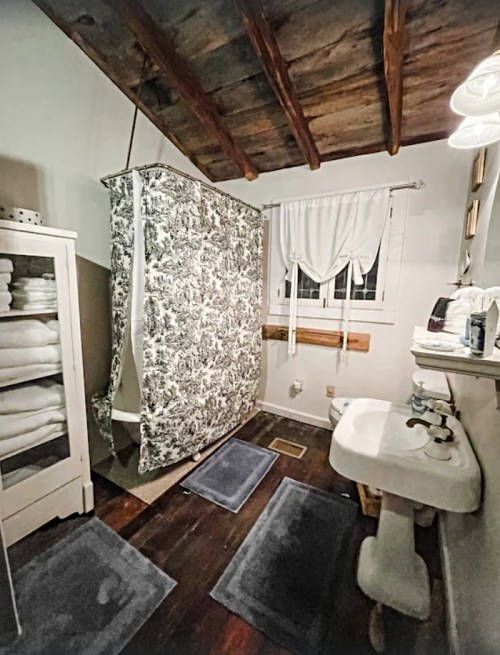 Cozy bathroom with a shower curtain, sink, and neatly arranged towels on shelves, featuring wooden beams overhead.