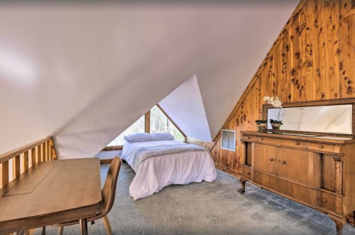 Cozy bedroom with a sloped ceiling, wooden accents, a bed, desk, and a vintage dresser near a large window.