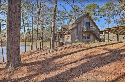 A rustic wooden cabin by a lake, surrounded by tall trees and a carpet of pine needles.
