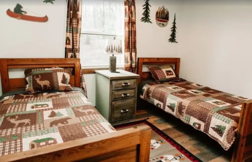Cozy bedroom with two wooden beds, quilted blankets, and rustic decor featuring trees and wildlife.