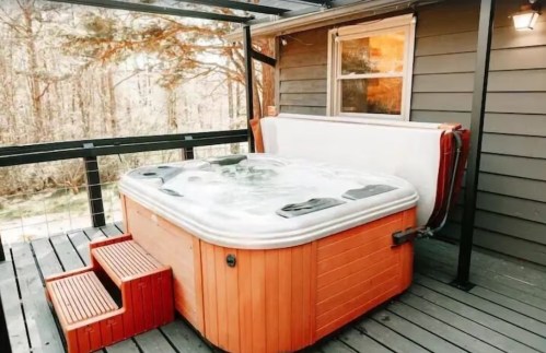A wooden deck features a hot tub with steps, surrounded by trees and a cozy cabin in the background.