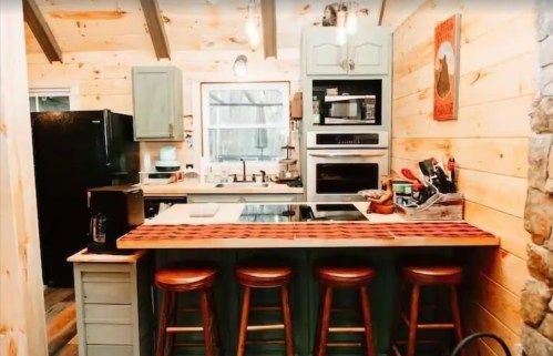 Cozy kitchen with wooden beams, green cabinets, a central island, and three wooden stools. Warm, rustic decor.