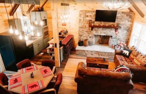 Cozy cabin interior featuring a stone fireplace, wooden furniture, and warm lighting, with a dining table and kitchen area.