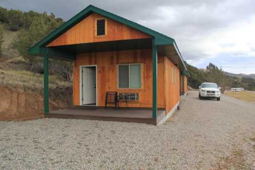 A small wooden cabin with a green roof, gravel driveway, and a porch with a chair, set in a rural landscape.