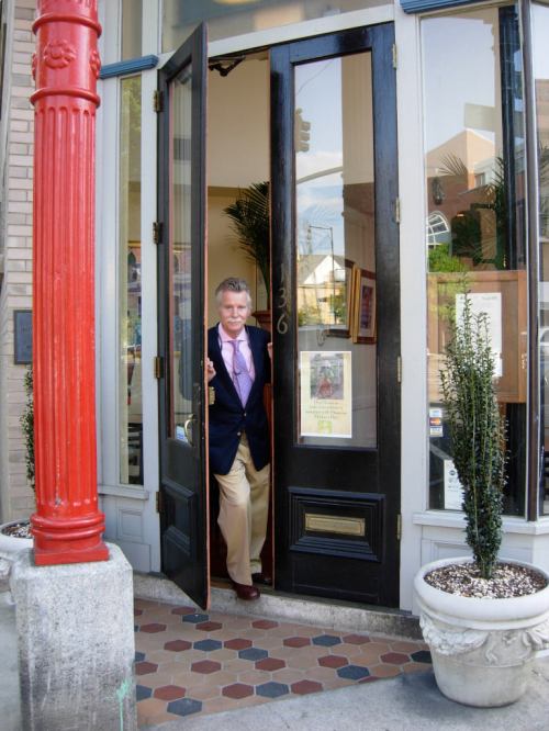 A man in a blazer stands at the entrance of a shop, holding the door open with a welcoming expression.