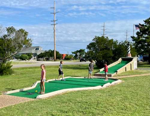 Four children play mini-golf on a sunny day, with a lighthouse and trees in the background.