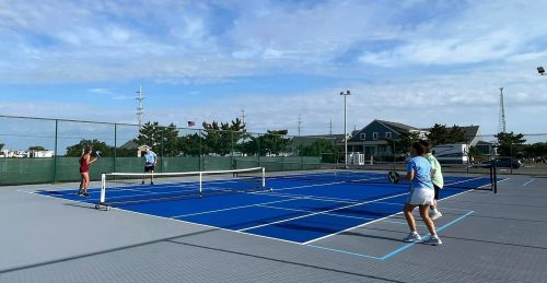 Two people playing tennis on a blue court, with a third person waiting to serve in the background. Clear sky above.