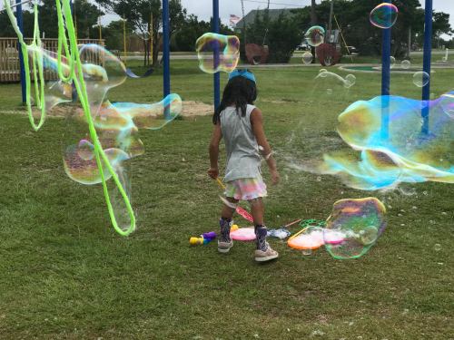 A child plays with colorful bubbles in a grassy park, surrounded by playground equipment.