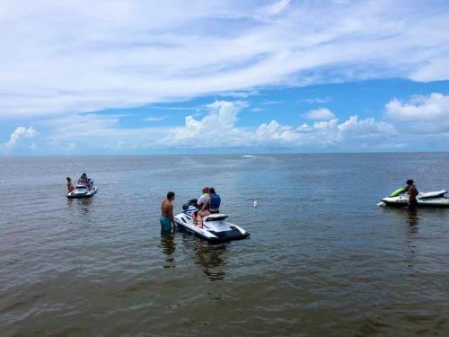 Two jet skis in calm water with people riding and standing on them, under a blue sky with scattered clouds.