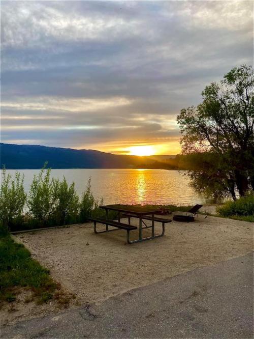 A serene lakeside view at sunset, featuring a picnic table and lush greenery along the water's edge.